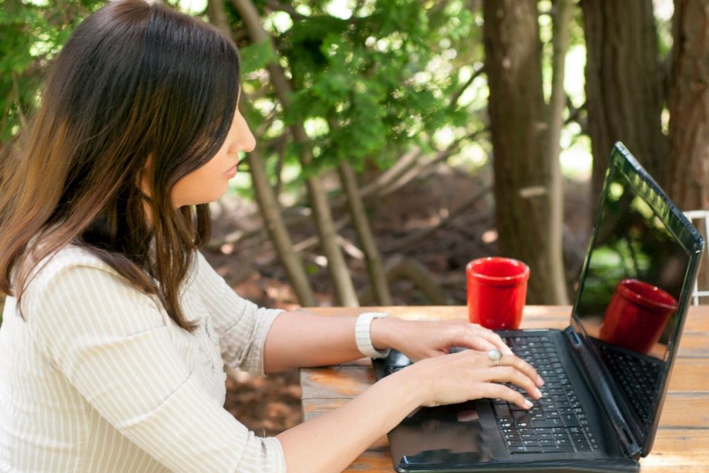 woman sits at picnic table with woods in the background, writing an article on her laptop
