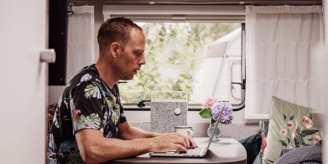 male freelance writer sits at RV table to work while surrounded by cozy home decor with a view of the outdoors over his shoulder